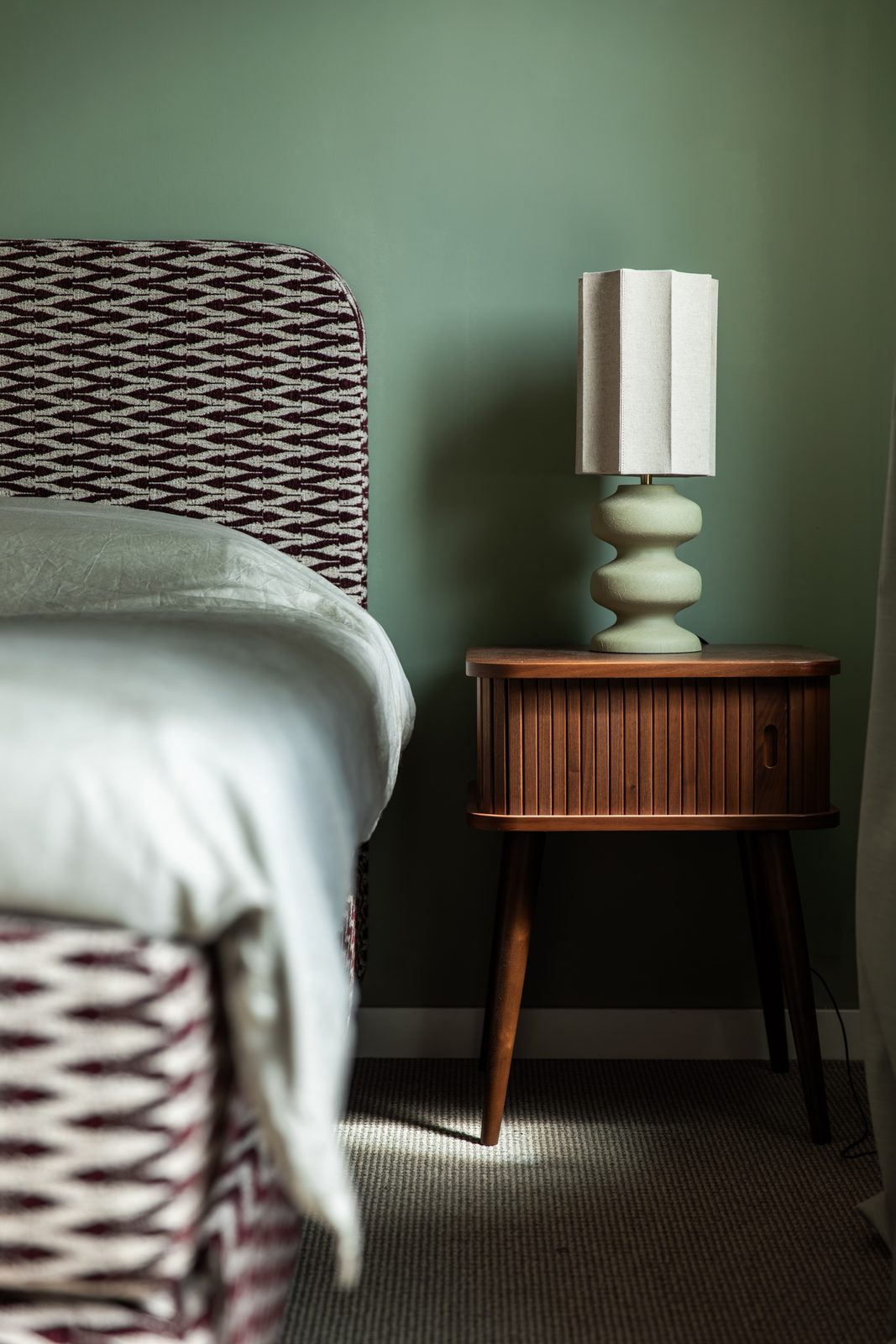 Hoofdbord met grafische stof tegen een groene wand, met een walnoten nachtkastje en keramieklamp, woning in Hillegom, ontwerp Authentic Interiors